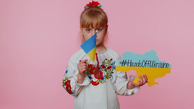 Upset toddler Ukrainian girl kid in embroidery dress protesting war conflict raises banner with inscription massage text on map Hands Off Ukraine on pink background. Peace, no war, stop aggression