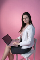 A girl on a pink background in a pink blouse, black skirt and high heels is sitting on a chair with a laptop on her lap. Programmer, freelancer, businesswoman or secretary