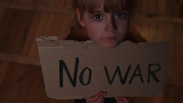 Close-up of afraid poor toddler girl kid homeless sitting at home floor at night holding banner with inscription massage No War. Child alone hiding from bombing attack. Crisis, peace, stop aggression