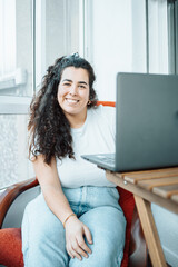 Happy plus size curly young woman smiling to camera while using a laptop to prepare,and do work...