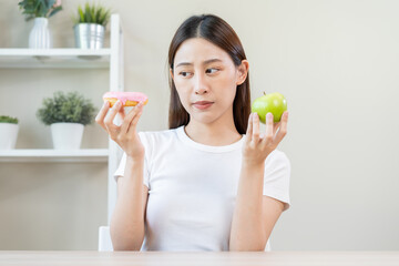 Diet, Dieting asian young woman or girl smile, confused choose, choice green apple or pink sweet donut, at home, eat food for good healthy, health when hungry. Close up female weight loss person.