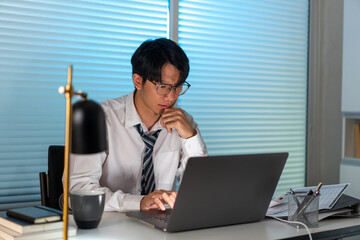 Overwork Concept The male who is wearing glasses resting his chin on his right hand and using his...