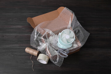 Small fan with bubble wrap in cardboard box and packaging items on dark wooden table, above view
