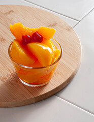 A glass full of slices of peach with syrup with two cherries on top, over a wooden choopin board, on a white surface kitchen. Vertical image.