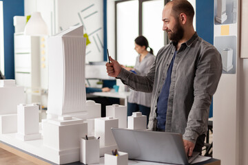 Inspired architect with laptop looking at the architectural lines of white foam model of residential project. Engineer holding pen inspecting the design of skyscraper maquette in modern office.