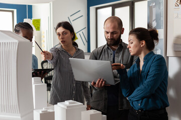 Couple of professional architects holding laptop looking at blueprints next to colleagues with tablet pointing at 3d maquette of skyscraper. Team of architectural engineers doing creative teamwork .
