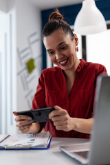 Employee in red shirt holding smartphone and looking at screen. Entrepreneur at the startup office in videocall conference. Businesswoman having a break playing casual mobile games online.