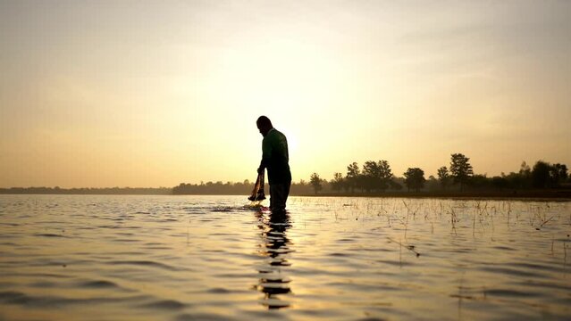Slow motion shot, Silhouette Asian Senior Fisherman wearing eyeglasses standing in water and clean fishing net after fishing at river in early morning