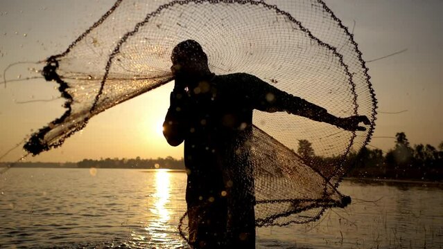 Slow motion and close up shot, Silhouette Asian Senior Fisherman wearing eyeglasses standing in water and prepairing fishing net after fishing at river in early morning
