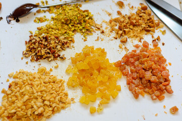 Heaps of candied fruits and nuts lie on a white cutting board, soft focus, a spoon in liquid chocolate lies on the background in defocus