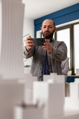 Architect using smartphone to record video of architectural maquette of skyscraper to post on social media. Bearded engineer photographing 3d white foam model with smart phone in modern office.