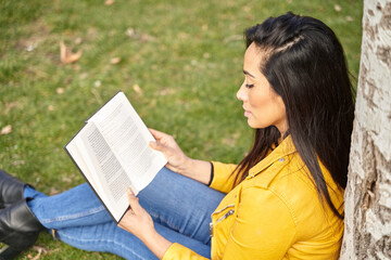 Side view of thoughtful Hispanic female reading novel while sitting with book under tree in summer garden