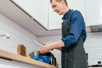 Portrait of middle-aged man wearing blue long-sleeve shirt and black apron breaking egg with knife into blue plastic round bowl on table in kitchen. Cooking, healthy food, diet, breakfast concept.