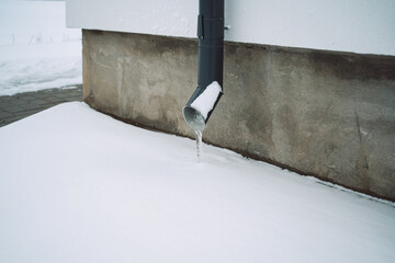 drain pipe with frozen water on the white wall of the house and snow around