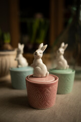 candles in ceramic containers top view. ceramic candlestick with a lid in the shape of a rabbit. decorative candles on the table near potted plants. ceramic figurine of a bunny	
