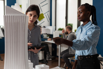 African american woman architect pointing pencil at skyscraper architectural foam model standing next to colleague engineer holding tablet in modern office. Architects doing teamwork.