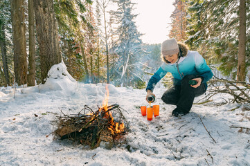 A woman at a campfire in winter in the forest pours tea from a thermos into glasses.