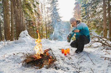 A woman at a campfire in winter in the forest pours tea from a thermos into glasses. - 488937588