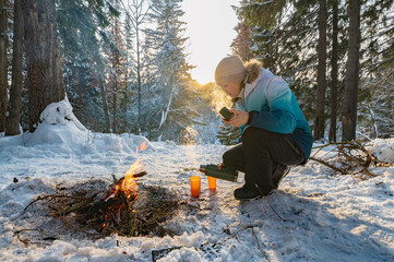 A woman at a campfire in winter in the forest pours tea from a thermos into glasses. - 488937589