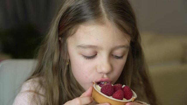 Adorable little girl eating fresh sweet raspberry cake indoors. Girl eating cake with cream and fruits. Child celebrates birthday with fresh raspberry tart
