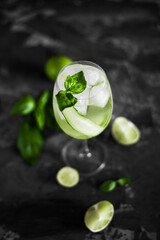 A wine glass of fresh cold coctail with ice, cucumber slices, lime fruit and basil leaves on the dark background