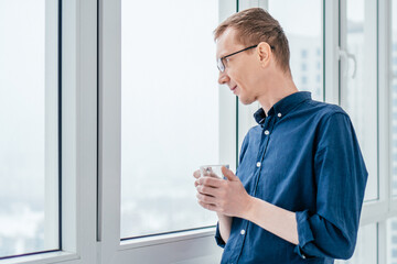 Portrait of focused middle-aged thin man with short ginger hair and glasses in blue long-sleeve shirt looking out through window thoughtfully, holding glass cup in bright sunny morning on balcony.