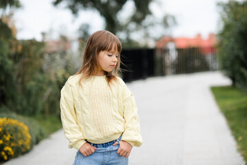 Fototapeta premium Stylish little kid girl 4-5 year old wear casual yellow knit woolen sweater and denim pants walk in park outdoors over nature background. Spring season. Springtime.