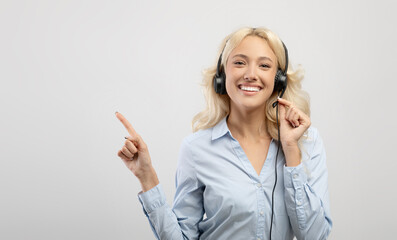 Happy young female call center operator wearing headset and pointing aside at free space, light background, panorama