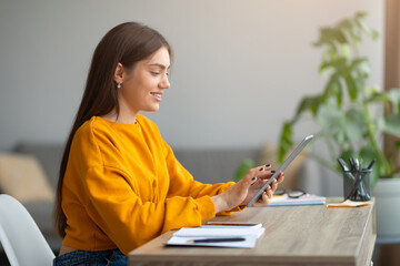 Side view of smiling young woman with digital tablet sitting at desk, working online from home...