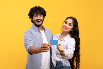 Happy indian couple with credit card promoting bank services, encouraging contactless shopping on yellow background
