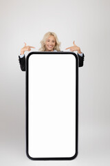 Young businesswoman standing behind huge smartphone and pointing at white empty screen, showing...