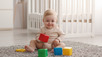 Portrait of adorable little baby smiling to camera, playing with colorful cubes, resting on floor at nursery, panorama