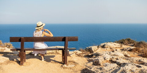 A stylish woman traveler watches a beautiful view of sea on the rocks on the beach. Summer vacation in Cyprus. Banner with copy space.