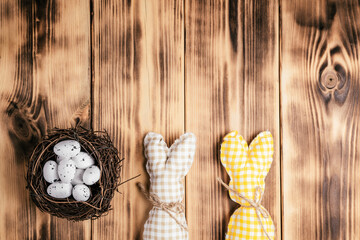 Composition of two handmade square brown, white and yellow colored bunnies with string-scarf on wooden natural background and white spot quail eggs in bird nest. Celebration easter holiday.