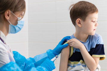 A young female doctor in a medical gown and mask inoculates a boy with a syringe against...