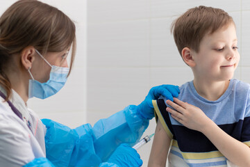 A young female doctor in a medical gown and mask inoculates a boy with a syringe against...