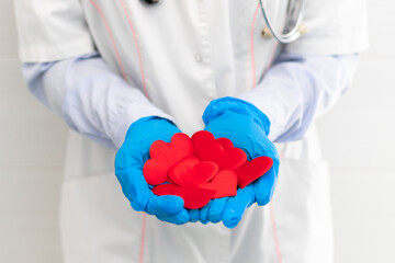 The hands of a doctor in a white medical coat and rubber gloves with a phonendoscope hold a lot of red hearts in the hospital. Selective focus. Close-up