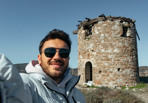 Happy handsome man with sun glasses takes a selfie portrait on vacation outdoor.  He is standing on old windmill. Travel alone concept.
