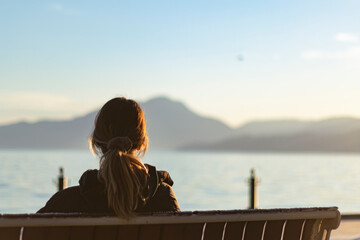 Back view of young woman sitting on bench in front of lake. Mountains silhouette at back. She has no friends. Loneliness concept. 