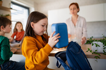 Little girl packing lunch box to backpack in kitchen at home.