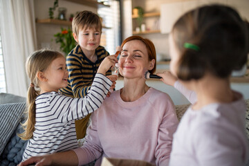 Three litttle children doing massage and apllying make up to their mother at home.