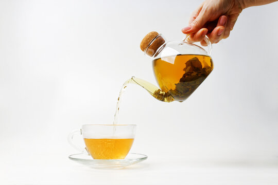 Human hand pours tea from a transparent kettle into a transparent cup on white background.