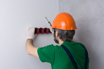 An electrician installs sockets in the apartment. A guy in an orange helmet and overalls makes electrics in the house.