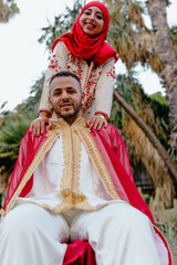 Obraz premium shot of head scarf dressed woman leaning on the shoulders of a muslim man while both wear traditional clothes and are in a place with palm trees