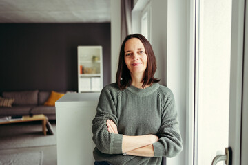 Portrait of smiling woman with arms crossed leaning by window at home