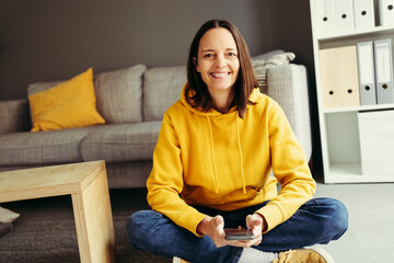 Portrait of smiling woman with smart phone sitting cross-legged against sofa at home