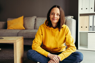 Portrait of woman wearing yellow hood sitting cross-legged against sofa at home