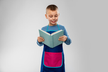 cooking, culinary and profession concept - happy smiling little boy in apron reading cook book over grey background