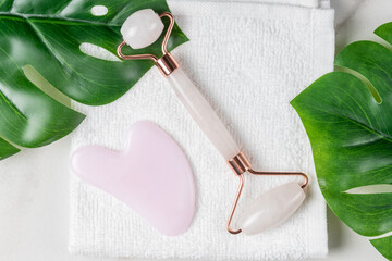 Gua Sha massager and face roller on white towel background with monstera leaves. Massage tools for facial skin care, SPA beauty treatment concept