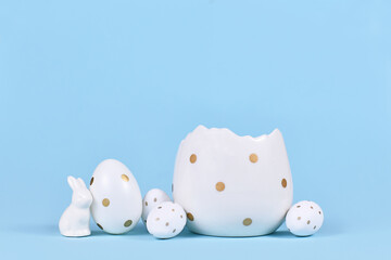 White easter eggs with golden dots, bunny figure and egg cup on blue background with copy space